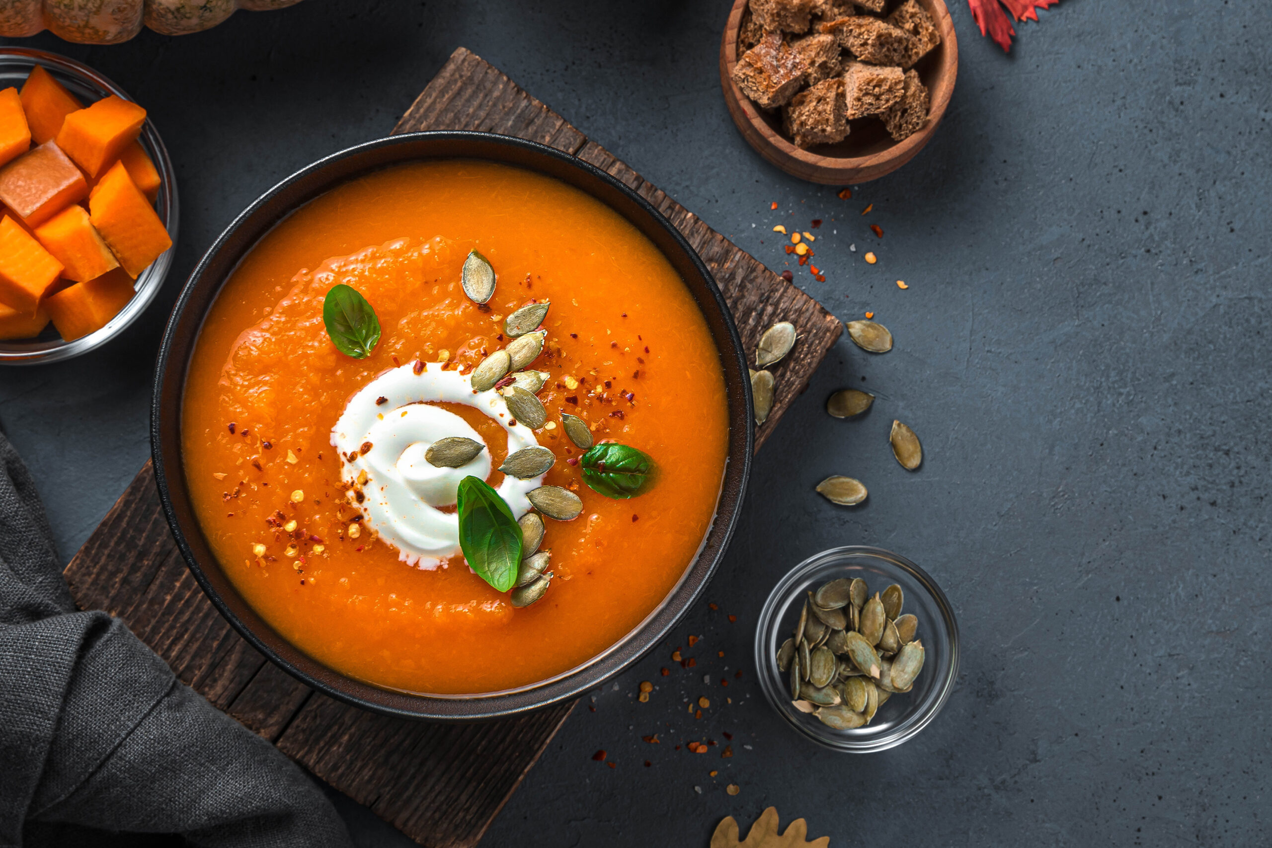 Eine Schüssel Kürbissuppe mit Sahne, Kürbiskernen und frischen Kräutern, umgeben von Kürbiswürfeln und Knäckebrot.